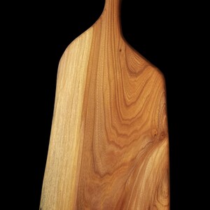May include: A large, wooden cutting board with a natural wood finish and a unique, asymmetrical shape. The board has a handle with a hole in the top.