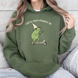 May include: Military green hoodie with a frog graphic riding a scooter and wearing a wizard hat. The text "Off to start chapter 30" is above the frog. The hoodie has a drawstring hood and a front pocket.