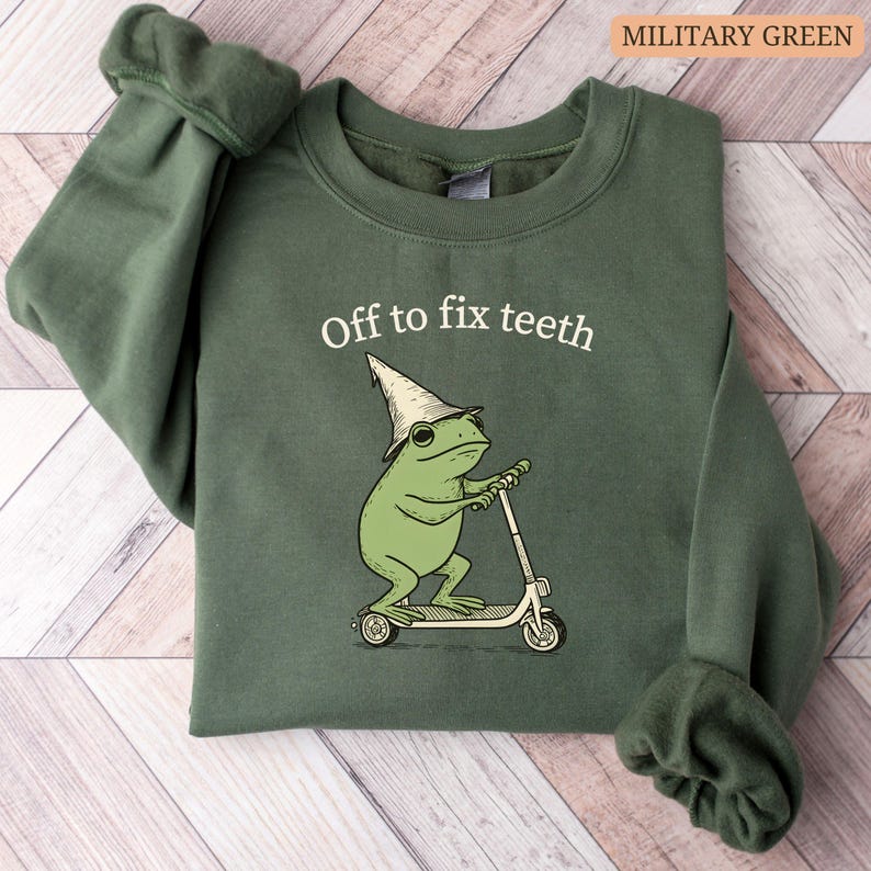 May include: Military green sweatshirt with the text "Off to fix teeth." The graphic features a green frog wearing a hat and riding a scooter. The sweatshirt is made of a soft material.