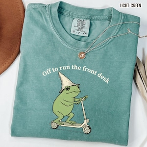K&ouml;nnte beinhalten: Hellgr&uuml;nes Comfort Colors T-Shirt mit dem Text "Off to run the front desk". Die Grafik zeigt einen gr&uuml;nen Frosch mit Zaubererhut, der einen Roller f&auml;hrt. Eine silberne Halskette mit Anh&auml;nger ist ebenfalls zu sehen.