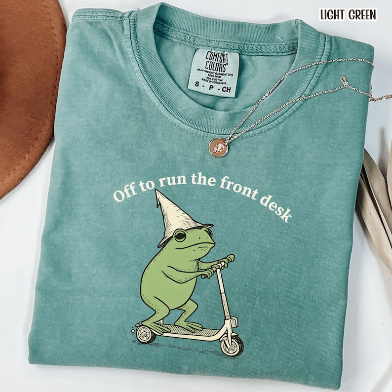 K&ouml;nnte beinhalten: Hellgr&uuml;nes Comfort Colors T-Shirt mit dem Text "Off to run the front desk". Die Grafik zeigt einen gr&uuml;nen Frosch mit Zaubererhut, der einen Roller f&auml;hrt. Eine silberne Halskette mit Anh&auml;nger ist ebenfalls zu sehen.