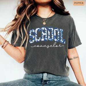 May include: Dark gray t-shirt with the words "SCHOOL counselor" in a blue and white floral design. The word "SCHOOL" is in a bold, collegiate-style font, while "counselor" is in a delicate script.