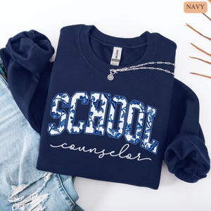 May include: Navy blue sweatshirt with the words "SCHOOL counselor" in white script. The word "SCHOOL" is in a bold, block letter style with a blue and white floral pattern. A silver necklace with a pendant is on the sweatshirt.