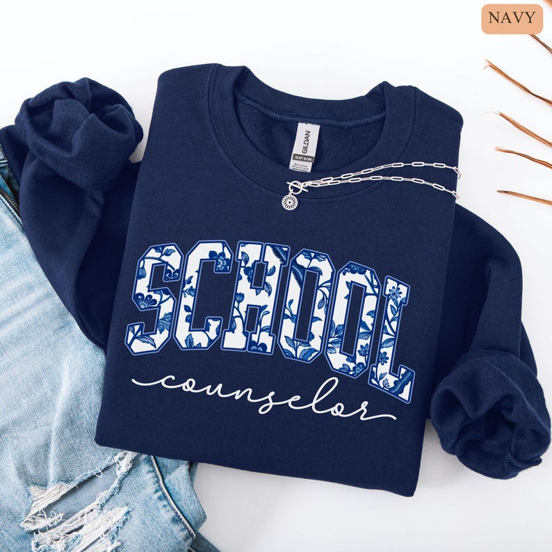 May include: Navy blue sweatshirt with the words "SCHOOL counselor" in white script. The word "SCHOOL" is in a bold, block letter style with a blue and white floral pattern. A silver necklace with a pendant is on the sweatshirt.
