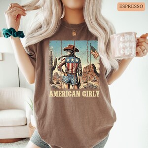 Puede incluir: Camiseta color espresso con una escena des&eacute;rtica que presenta a una vaquera con sombrero y pantalones cortos de mezclilla, con una bandera estadounidense de fondo. El texto "AMERICAN GIRLY" est&aacute; impreso debajo.