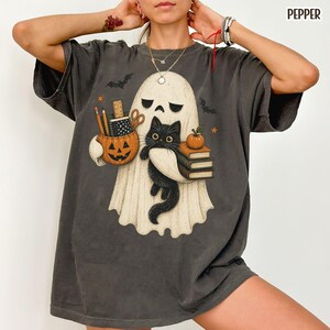 May include: A charcoal gray short-sleeve t-shirt featuring a graphic of a white ghost holding a black cat, a stack of books, and a jack-o'-lantern bucket filled with school supplies. The design includes small bats and stars, with the word PEPPER in the corner.