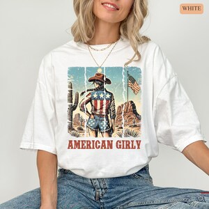 Puede incluir: Camiseta blanca con un dise&ntilde;o de una vaquera con atuendo patri&oacute;tico americano, un paisaje des&eacute;rtico y la bandera estadounidense. El texto "AMERICAN GIRLY" est&aacute; debajo. La camiseta tiene un corte holgado y de gran tama&ntilde;o.