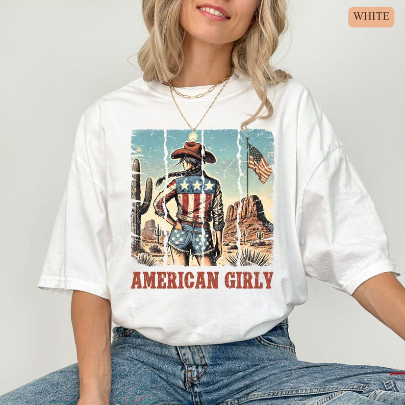 Puede incluir: Camiseta blanca con un dise&ntilde;o de una vaquera con atuendo patri&oacute;tico americano, un paisaje des&eacute;rtico y la bandera estadounidense. El texto "AMERICAN GIRLY" est&aacute; debajo. La camiseta tiene un corte holgado y de gran tama&ntilde;o.