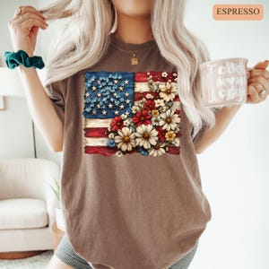 May include: Espresso-colored t-shirt featuring a distressed American flag design. The flag's stars are replaced with blue flowers, and the stripes are adorned with red, white, and yellow flowers. The shirt has a relaxed fit.