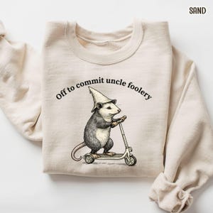May include: Sand-colored sweatshirt with a graphic of an opossum wearing a wizard hat and riding a scooter. The text "Off to commit uncle foolery" is arched above the opossum. The sweatshirt is made of a soft material.