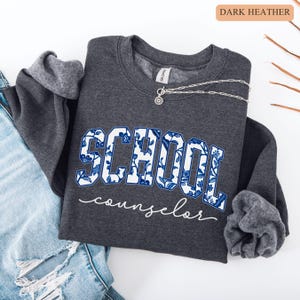 May include: Dark heather gray sweatshirt with the word "SCHOOL" in a blue floral design and the word "counselor" in white script. A silver necklace with a pendant is draped across the sweatshirt. The sweatshirt is folded on top of a pair of blue jeans.
