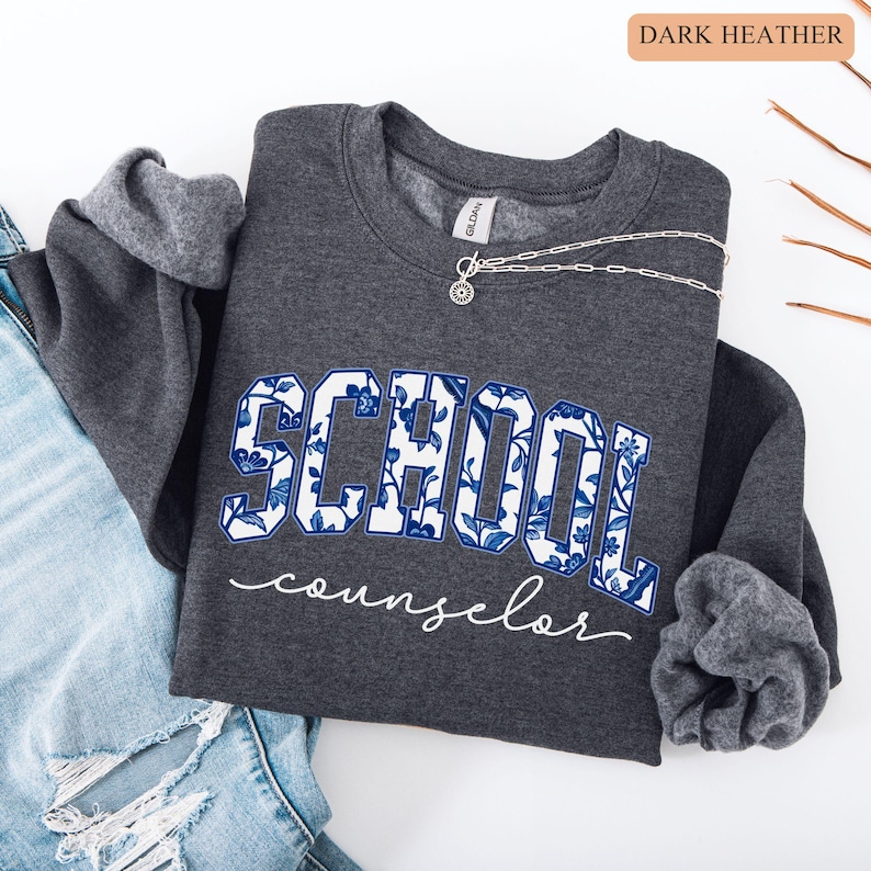 May include: Dark heather gray sweatshirt with the word "SCHOOL" in a blue floral design and the word "counselor" in white script. A silver necklace with a pendant is draped across the sweatshirt. The sweatshirt is folded on top of a pair of blue jeans.