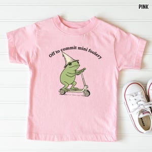 Puede incluir: Camiseta rosa claro para ni&ntilde;os con la frase "Off to commit mini foolery" y un dibujo de una rana en un patinete, con un sombrero de mago. Zapatillas blancas visibles en la esquina inferior derecha.