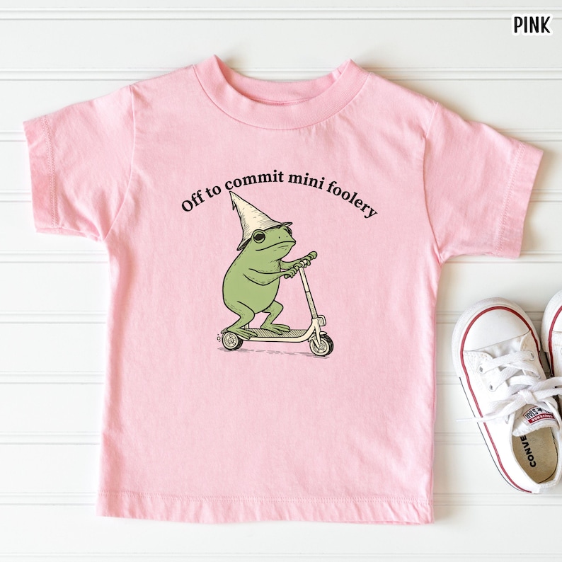 Puede incluir: Camiseta rosa claro para ni&ntilde;os con la frase "Off to commit mini foolery" y un dibujo de una rana en un patinete, con un sombrero de mago. Zapatillas blancas visibles en la esquina inferior derecha.