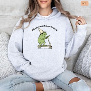 May include: Light gray hoodie with the text "Off to commit mom foolery." The graphic features a green frog wearing a wizard hat and riding a scooter. The hoodie is made of a soft material and has a drawstring hood.