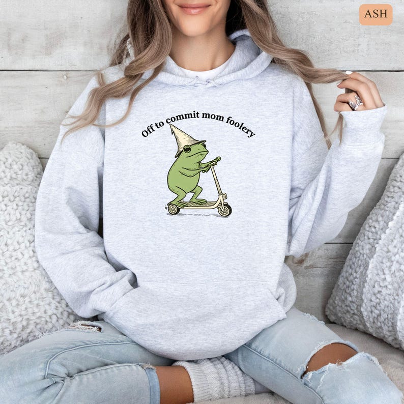 May include: Light gray hoodie with the text "Off to commit mom foolery." The graphic features a green frog wearing a wizard hat and riding a scooter. The hoodie is made of a soft material and has a drawstring hood.