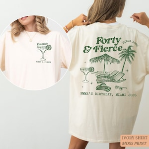 Può includere: T-shirt color avorio con grafica verde muschio. Il davanti presenta un cocktail e il testo "Emma's" e "Forty & Fierce". Il retro mostra "Forty & Fierce Since 1986" con una scena di spiaggia e "Emma's Birthday, Miami 2026".