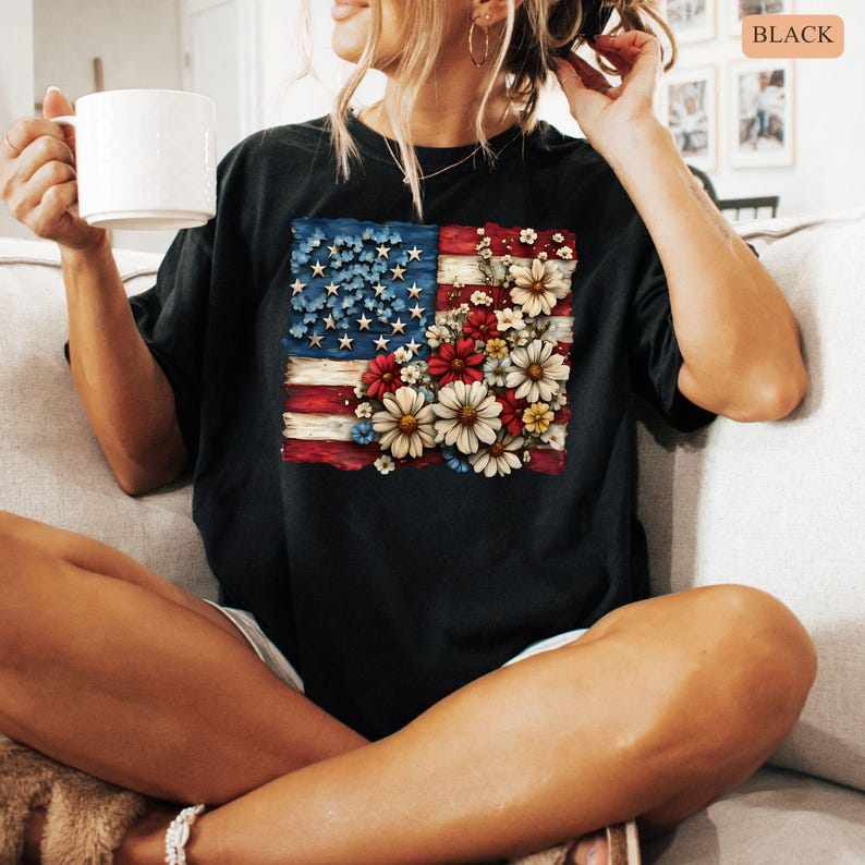 May include: Black t-shirt featuring a floral American flag design. The flag incorporates red and white stripes with blue stars and is adorned with colorful flowers. The shirt is a casual style.