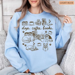May include: Light blue sweatshirt with a graphic design featuring dogs, coffee cups, books, and paw prints. The text "dogs. coffee. books." is printed at the top. The design includes illustrations of dogs, books, and coffee cups.