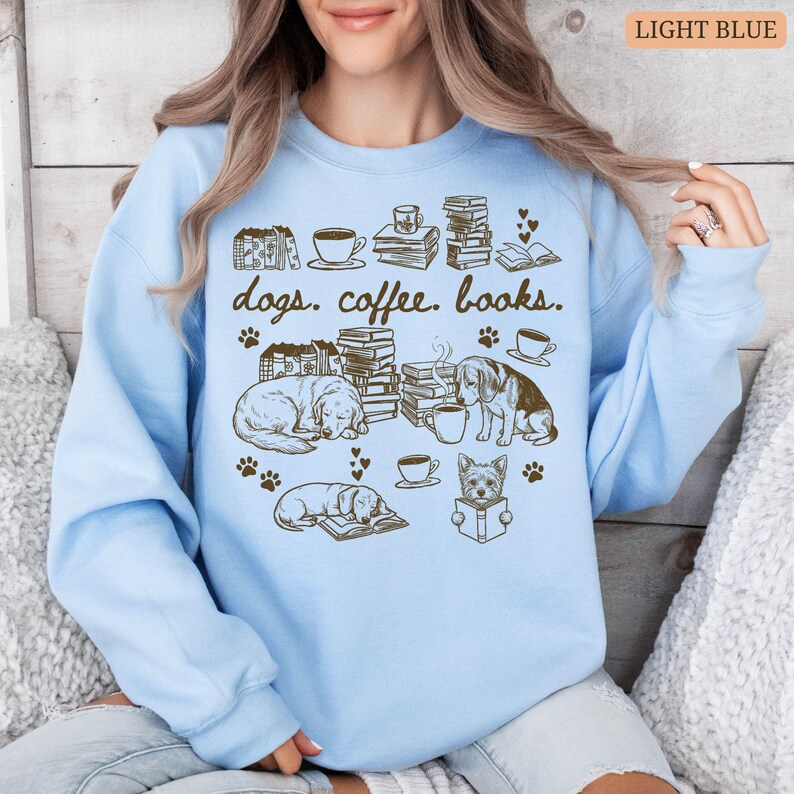 May include: Light blue sweatshirt with a graphic design featuring dogs, coffee cups, books, and paw prints. The text "dogs. coffee. books." is printed at the top. The design includes illustrations of dogs, books, and coffee cups.