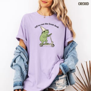 K&ouml;nnte beinhalten: Ein helllila T-Shirt mit dem Text "Off to run the front desk". Das Design zeigt einen Cartoon-Frosch mit Zaubererhut auf einem Roller. Das Wort "ORCHID" ist oben rechts aufgedruckt.