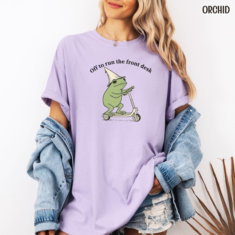 K&ouml;nnte beinhalten: Ein helllila T-Shirt mit dem Text "Off to run the front desk". Das Design zeigt einen Cartoon-Frosch mit Zaubererhut auf einem Roller. Das Wort "ORCHID" ist oben rechts aufgedruckt.