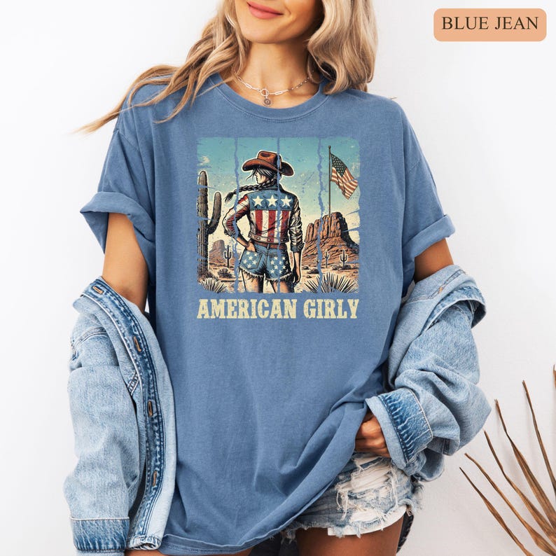 Puede incluir: Camiseta azul vaquero con un gr&aacute;fico de una vaquera en un paisaje des&eacute;rtico, con sombrero y ropa con dise&ntilde;o de bandera americana. El texto "AMERICAN GIRLY" est&aacute; impreso debajo.