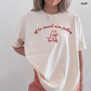 May include: An off-white short-sleeve t-shirt featuring a red graphic of a frog riding a scooter. The text "off to commit mom foolery" is printed in a red cursive font above the frog illustration. The shirt has a crew neckline and a relaxed fit.