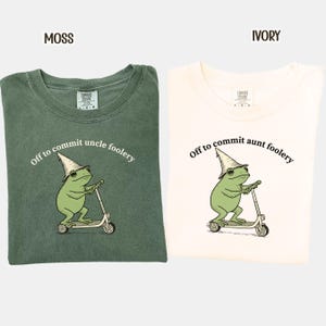 May include: Two t-shirts, one moss green and one ivory, each with a graphic of a frog riding a scooter and wearing a wizard hat. The moss green shirt reads "Off to commit uncle foolery," and the ivory shirt reads "Off to commit aunt foolery."