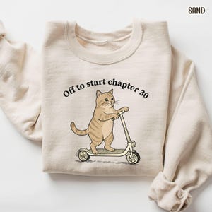 May include: Sand-colored sweatshirt with the text "Off to start chapter 30." A cartoon cat is riding a scooter. The cat is tan with brown stripes. The scooter is white and black.