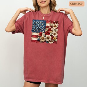 May include: Crimson-colored t-shirt featuring a distressed American flag design. The flag is partially replaced with a floral arrangement of red, white, and blue flowers. The shirt has short sleeves and a crew neck.