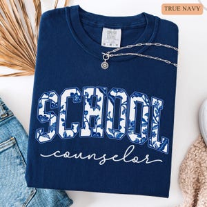 May include: A navy blue sweatshirt with the words "SCHOOL counselor" in a white floral design. The sweatshirt has a crew neck and is paired with a silver necklace. The text "TRUE NAVY" is in the upper right corner.
