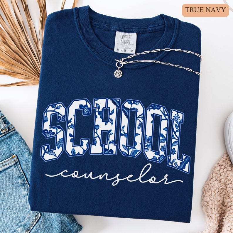 May include: A navy blue sweatshirt with the words "SCHOOL counselor" in a white floral design. The sweatshirt has a crew neck and is paired with a silver necklace. The text "TRUE NAVY" is in the upper right corner.