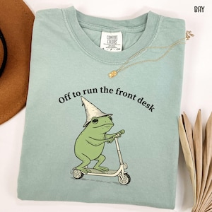 K&ouml;nnte beinhalten: Hellgr&uuml;nes Comfort Colors T-Shirt mit dem Text "Off to run the front desk". Das Shirt zeigt einen Cartoon-Frosch mit Zaubererhut, der einen Roller f&auml;hrt. Eine goldene Halskette ist &uuml;ber dem Shirt drapiert.