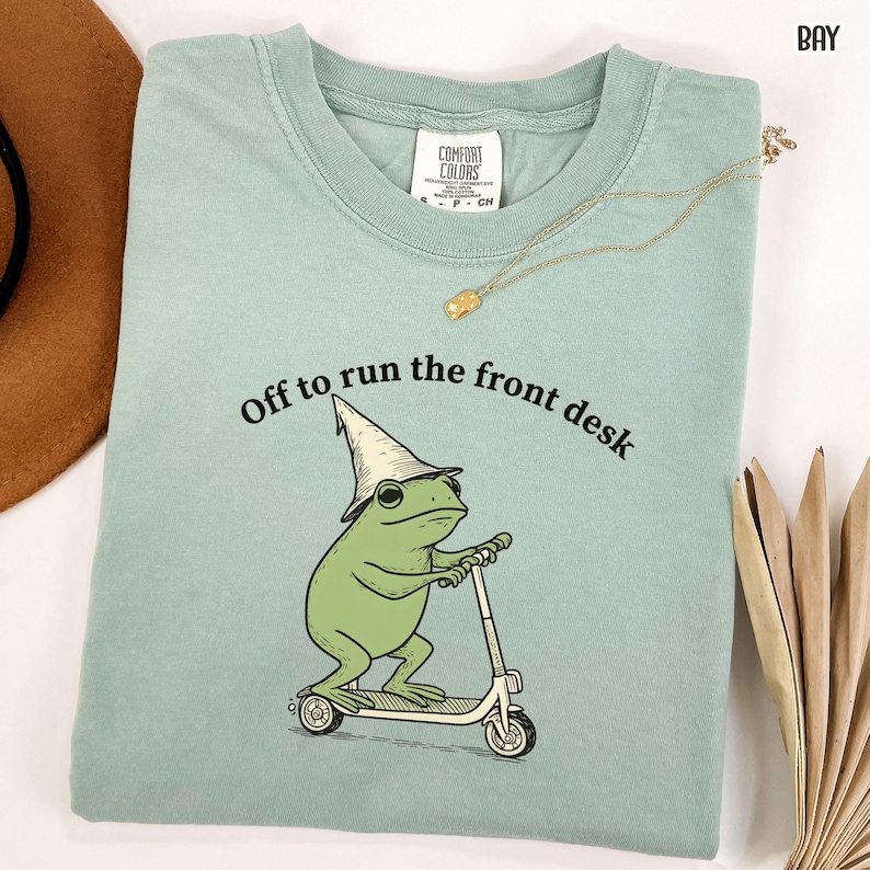 K&ouml;nnte beinhalten: Hellgr&uuml;nes Comfort Colors T-Shirt mit dem Text "Off to run the front desk". Das Shirt zeigt einen Cartoon-Frosch mit Zaubererhut, der einen Roller f&auml;hrt. Eine goldene Halskette ist &uuml;ber dem Shirt drapiert.