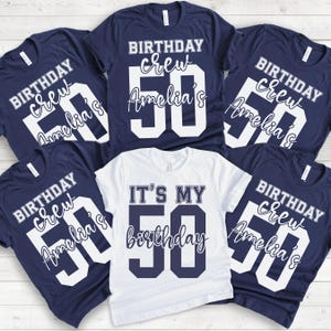 May include: A collection of five t-shirts. Four navy blue shirts feature white text reading BIRTHDAY crew Amelia's 50. One white shirt in the center features navy blue text reading IT'S MY 50 birthday. The shirts are arranged in a group display.