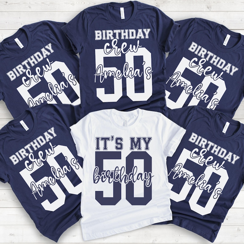 May include: A collection of five t-shirts. Four navy blue shirts feature white text reading BIRTHDAY crew Amelia's 50. One white shirt in the center features navy blue text reading IT'S MY 50 birthday. The shirts are arranged in a group display.