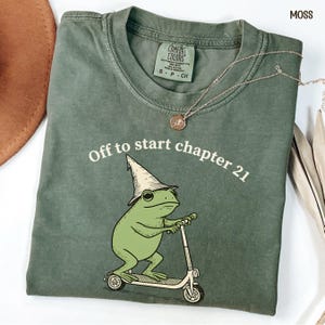 May include: Moss green t-shirt with the text "Off to start chapter 21" and a cartoon frog wearing a wizard hat and riding a scooter. A silver necklace with a pendant is on the shirt.