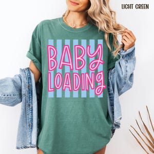 May include: Light green t-shirt with the text "BABY LOADING" in pink letters outlined in white, set against a light blue striped background. The shirt has short sleeves and a relaxed fit.