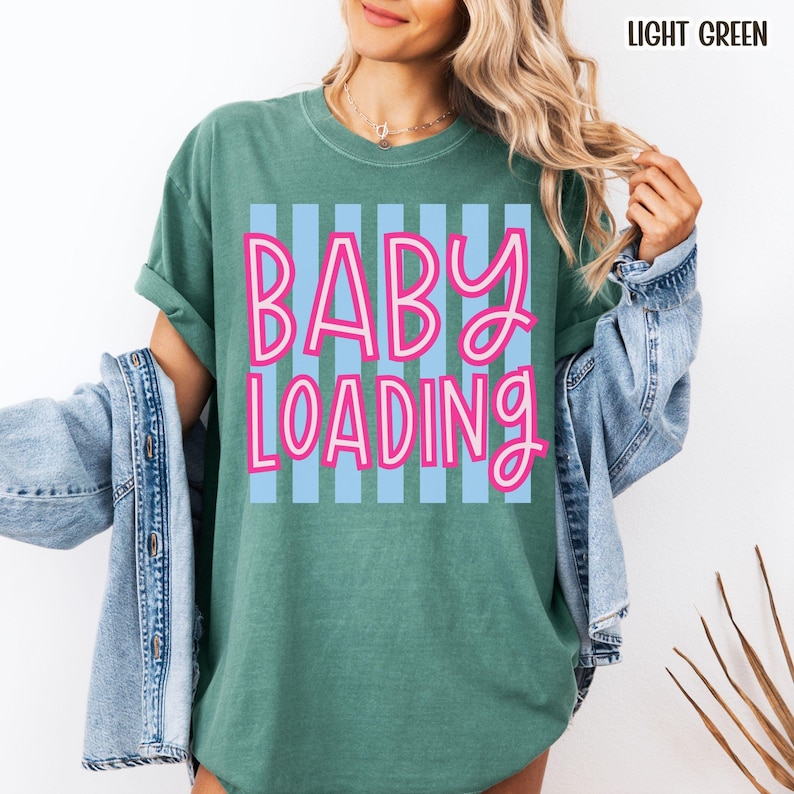 May include: Light green t-shirt with the text "BABY LOADING" in pink letters outlined in white, set against a light blue striped background. The shirt has short sleeves and a relaxed fit.