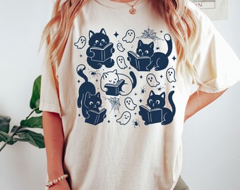 May include: Ivory t-shirt with a navy blue graphic of cats reading books, ghosts, spiders, and cobwebs. The design has a Halloween theme. The shirt is a relaxed fit, suitable for casual wear.