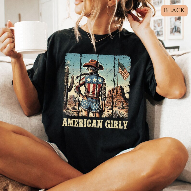 Puede incluir: Camiseta negra con un gr&aacute;fico de una vaquera en un paisaje des&eacute;rtico, con una chaqueta y pantalones cortos con un dise&ntilde;o de bandera estadounidense. El texto "AMERICAN GIRLY" est&aacute; impreso debajo.