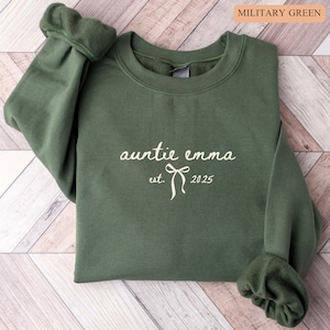 May include: Military green sweatshirt with the text "auntie emma est. 2025" in a cursive font. The sweatshirt has a crew neck and long sleeves with ribbed cuffs. The background is a light wood grain pattern.