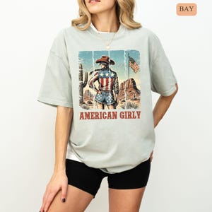 Puede incluir: Camiseta verde claro con un gr&aacute;fico de una persona con sombrero de vaquero y pantalones cortos de mezclilla, con una bandera estadounidense y un paisaje des&eacute;rtico. El texto "AMERICAN GIRLY" est&aacute; impreso debajo.