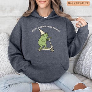 May include: Dark heather gray hoodie with a front graphic of a green frog riding a scooter and wearing a wizard hat. The text "Off to commit mom foolery" is above the frog. The hoodie has a drawstring hood and a front pocket.