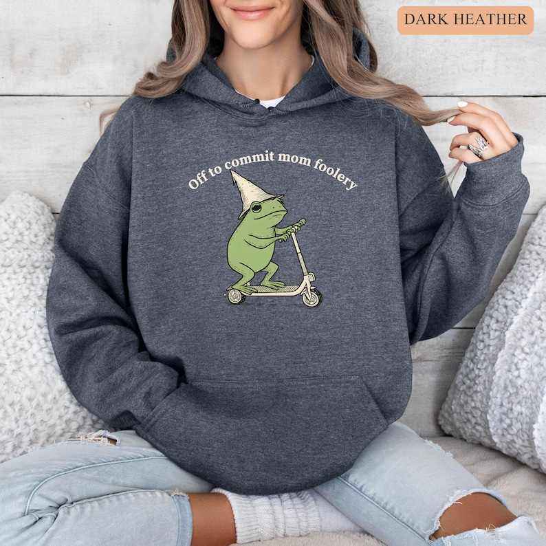 May include: Dark heather gray hoodie with a front graphic of a green frog riding a scooter and wearing a wizard hat. The text "Off to commit mom foolery" is above the frog. The hoodie has a drawstring hood and a front pocket.