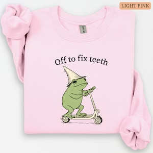 May include: Light pink sweatshirt with the text "Off to fix teeth." The graphic shows a green frog wearing a hat and riding a scooter. The sweatshirt is a solid light pink color.