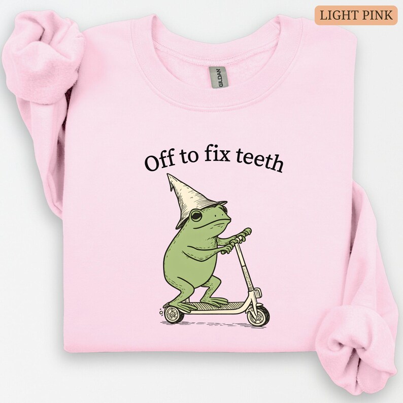 May include: Light pink sweatshirt with the text "Off to fix teeth." The graphic shows a green frog wearing a hat and riding a scooter. The sweatshirt is a solid light pink color.
