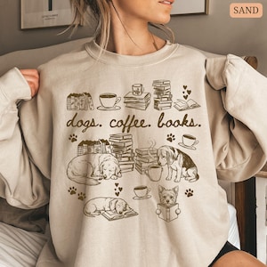 May include: Sand-colored sweatshirt with a graphic featuring dogs, coffee cups, books, and the text "dogs. coffee. books." The design includes illustrations of sleeping dogs, stacks of books, and coffee cups. Paw prints and hearts are also included in the design.