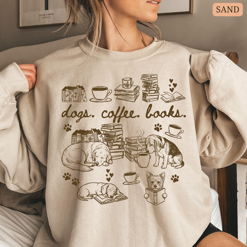 May include: Sand-colored sweatshirt with a graphic featuring dogs, coffee cups, books, and the text "dogs. coffee. books." The design includes illustrations of sleeping dogs, stacks of books, and coffee cups. Paw prints and hearts are also included in the design.